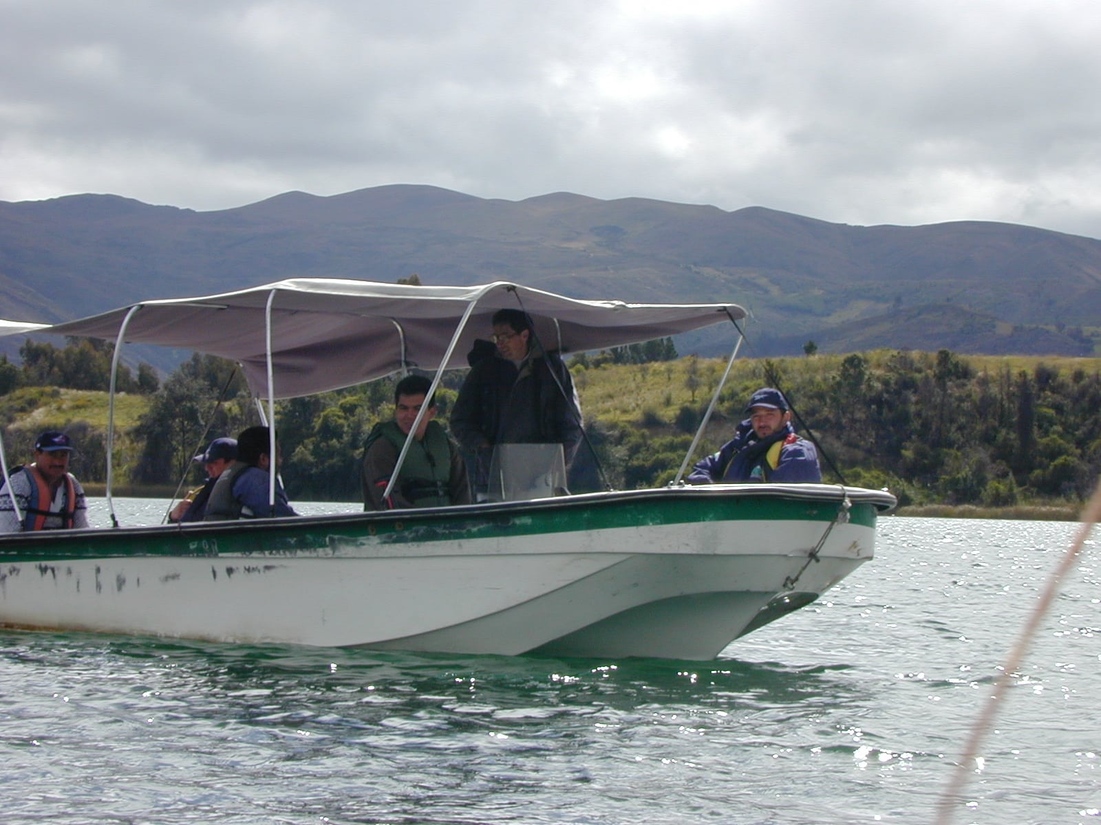 Five people sit on a covered boat in the water. Forests and mountains rise in the background.