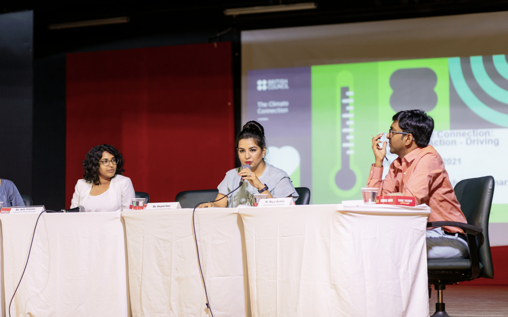 Three people sit at a table with microphones. Two are listening while one in the middle speaks in front of a presentation.