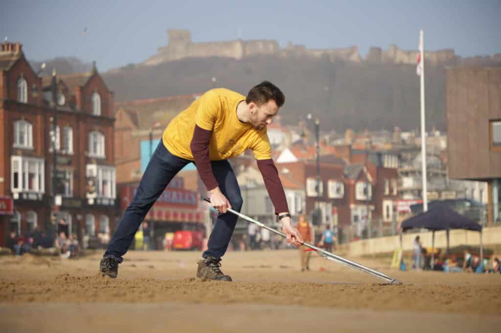 A person in a yellow team t-shirt rakes the sand to help create the art piece, with city buildings and beachgoers in the background.