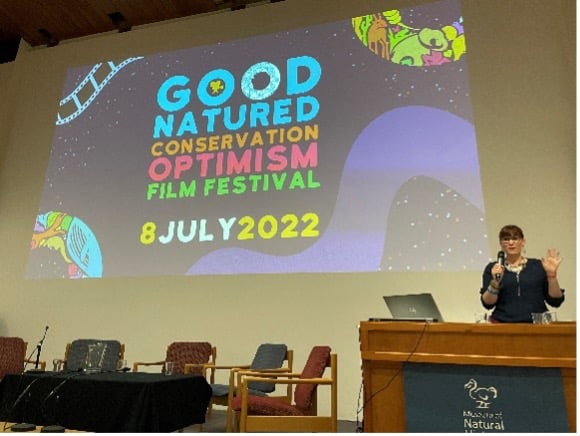 A person standing at a podium introduces the Good Natured Conservation Optimism Film Festival in front of a screen announcing the event on 8 July 2022.